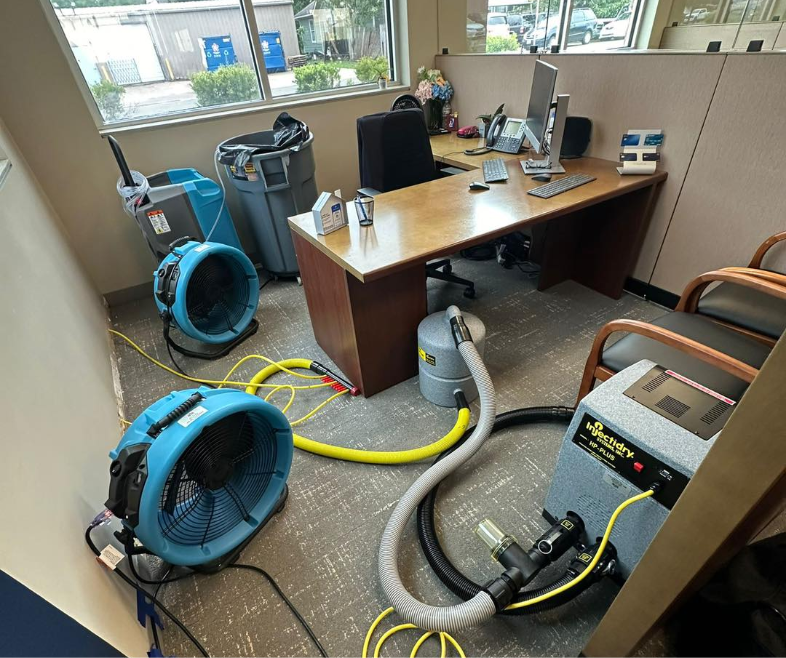 Interior of an office with an L-shaped desk and two chairs in front of it with industrial fans and other water restoration equipment on the floor